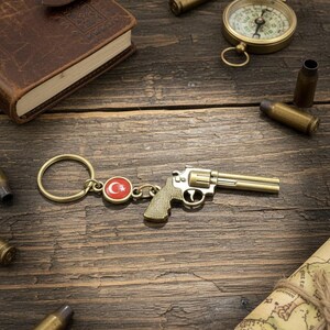 Könnte beinhalten: Ein goldfarbener Revolver-Schlüsselanhänger mit türkischem Flaggenemblem, auf einer rustikalen Holzoberfläche. Die Szene umfasst einen Kompass, ein ledergebundenes Notizbuch und mehrere Patronenhülsen, was auf ein Thema von Reisen oder Abenteuer hindeutet.