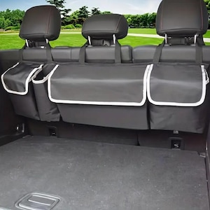 May include: Black car seat organizer with multiple storage compartments. Features a large central compartment and two side pockets, all trimmed with white piping. The organizer hangs from the headrests, providing convenient storage solutions for car interiors.