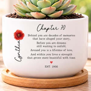 May include: A white ceramic planter with a bamboo saucer containing a succulent. The planter features a red poppy illustration, the name Cynthia, and the text: Chapter 70, Behind you are decades of memories that have shaped your story, Before you are dreams still waiting to unfold, Around you is a lifetime of love, And within you lives a strength that grows more beautiful with time. EST. 1956.