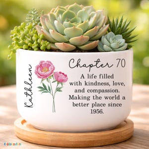 May include: White ceramic planter with a variety of succulents. The planter has the name "Kathleen" and the words "Chapter 70" with the text "A life filled with kindness, love, and compassion. Making the world a better place since 1956." Pink floral design.