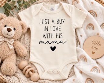 May include: A cream-colored short-sleeve baby bodysuit featuring the black printed text JUST A BOY IN LOVE WITH HIS mama and a small black heart icon centered on the front. The garment has a round neckline and three snaps at the bottom.