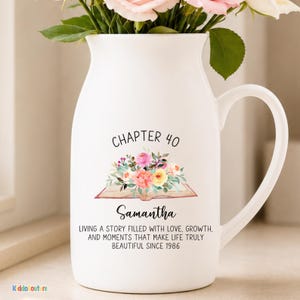 May include: White ceramic pitcher with a curved handle, holding pink and white flowers. The pitcher has the text "CHAPTER 40" and "Samantha" with a floral design. The text below reads "LIVING A STORY FILLED WITH LOVE, GROWTH, AND MOMENTS THAT MAKE LIFE TRULY BEAUTIFUL SINCE 1986."