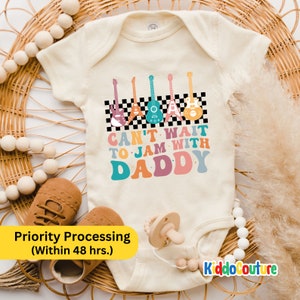 May include: A white baby onesie with a colorful graphic of guitars and the text "Can't wait to jam with Daddy".
