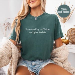 May include: Blue spruce-colored t-shirt with the text "Powered by caffeine and plot twists." The shirt is a short-sleeved, crew neck style. A circular graphic in the upper right corner indicates the color of the shirt. The model is wearing denim shorts and a cream-colored sweater.