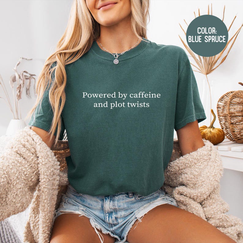 May include: Blue spruce-colored t-shirt with the text "Powered by caffeine and plot twists." The shirt is a short-sleeved, crew neck style. A circular graphic in the upper right corner indicates the color of the shirt. The model is wearing denim shorts and a cream-colored sweater.