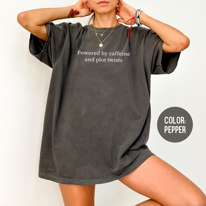 May include: Pepper-colored oversized t-shirt with the text "Powered by caffeine and plot twists" in white. The shirt has short sleeves and a relaxed fit. The color is indicated in a circular label.