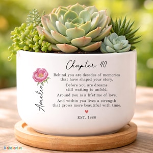 May include: A white ceramic planter with a succulent arrangement. The planter has the text "Chapter 40" and a poem. A pink floral design and the name "Amelia" are also featured. The planter sits on a wooden base.