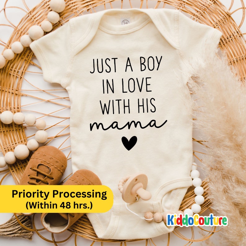 May include: A white baby onesie with the text "JUST A BOY IN LOVE WITH HIS mama" in black lettering with a small black heart below the text. The onesie is lying on a woven wicker surface with a wooden teether and brown baby shoes.