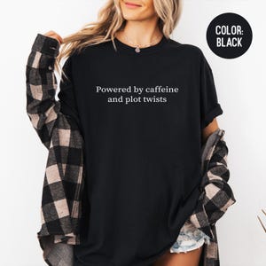 May include: Black t-shirt with the text "Powered by caffeine and plot twists" in white. The shirt is a solid black color. A black circle with the words "COLOR: BLACK" is in the upper right corner.