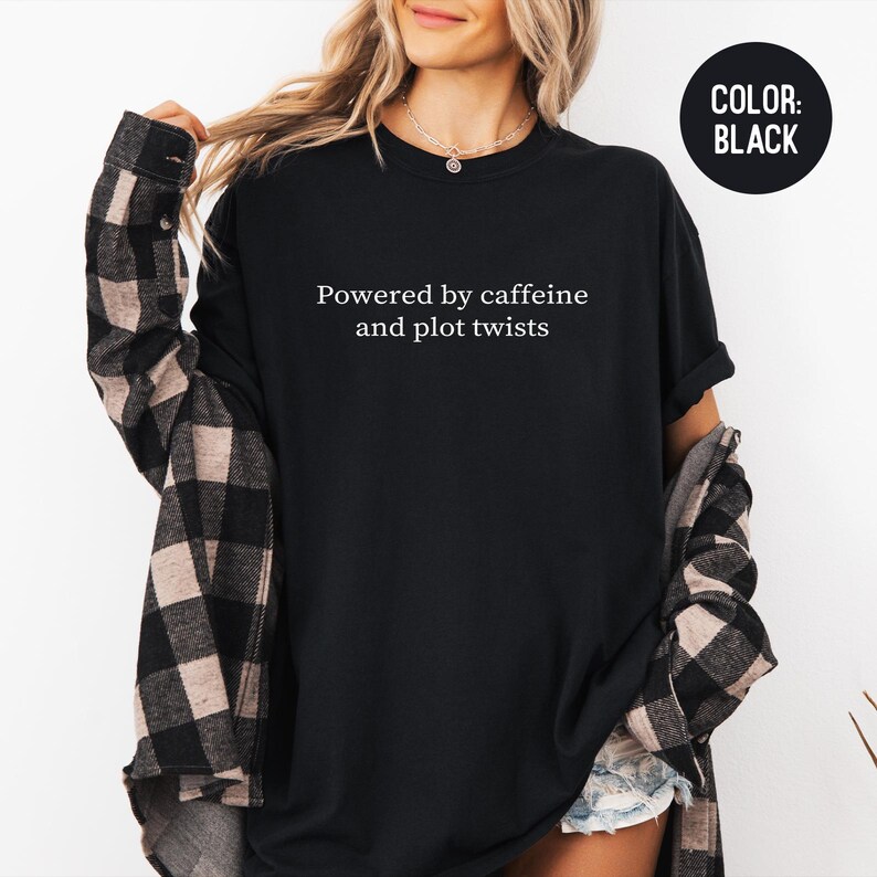 May include: Black t-shirt with the text "Powered by caffeine and plot twists" in white. The shirt is a solid black color. A black circle with the words "COLOR: BLACK" is in the upper right corner.