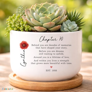 May include: A white ceramic planter with a variety of succulents. The planter has the text "Chapter 70" and a poem. A red flower and the name "Cynthia" are also on the planter. The planter sits on a wooden coaster.