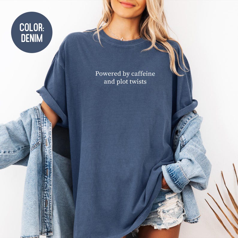 May include: Denim blue t-shirt with the text "Powered by caffeine and plot twists" in white. The shirt has rolled-up sleeves and is paired with a denim jacket and shorts. A circular label in the upper left corner reads "COLOR: DENIM".