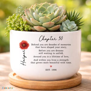May include: A white ceramic planter on a round wooden base containing various green succulents. The planter features a red poppy illustration, the name Harper, and text reading Chapter 30, a poem about memories and strength, and EST. 1996.