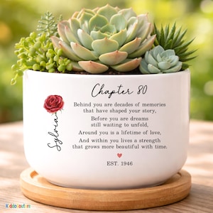 May include: A white ceramic planter with a succulent arrangement. The planter has the text "Chapter 80" and a poem. A red rose and the name "Sylvia" are also on the planter. The planter sits on a wooden saucer.