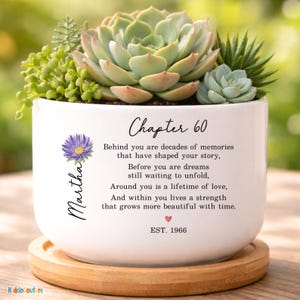 May include: A white ceramic planter with a variety of succulents. The planter has the text "Chapter 60" and a poem. A purple flower and the name "Martha" are also on the planter. The planter sits on a wooden coaster.