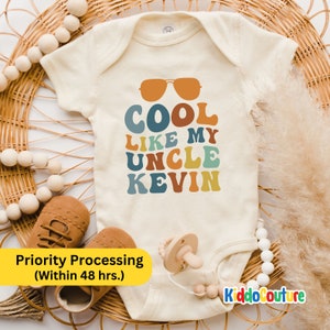 May include: A white baby onesie with a retro-style graphic that says "COOL LIKE MY UNCLE KEVIN" in colorful letters. The graphic also includes a pair of sunglasses.