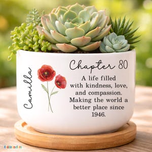 May include: White ceramic planter with a variety of succulents. The planter has the name "Camilla" and the text "Chapter 80" with the words "A life filled with kindness, love, and compassion. Making the world a better place since 1946."