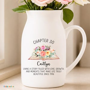 May include: White ceramic pitcher with a curved handle, holding pink and peach flowers. The pitcher has the text "CHAPTER 30" and "Caitlyn" with a floral design. The text below reads "LIVING A STORY FILLED WITH LOVE, GROWTH, AND MOMENTS THAT MAKE LIFE TRULY BEAUTIFUL SINCE 1996."