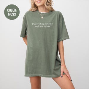 May include: Moss green oversized t-shirt with the text "Powered by caffeine and plot twists" in white. The shirt has short sleeves and a relaxed fit. The color is indicated by a circular label in the upper left corner.