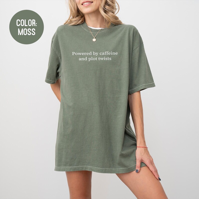 May include: Moss green oversized t-shirt with the text "Powered by caffeine and plot twists" in white. The shirt has short sleeves and a relaxed fit. The color is indicated by a circular label in the upper left corner.