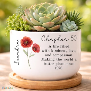 May include: A white ceramic planter with a variety of succulents and greenery. The planter features the text "Chapter 50" and "A life filled with kindness, love, and compassion. Making the world a better place since 1976." Includes a wooden saucer.