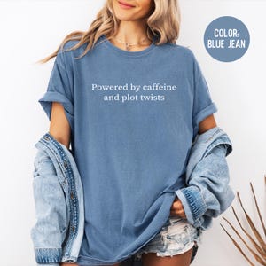 May include: A blue jean colored t-shirt with the text "Powered by caffeine and plot twists" in white. The shirt has rolled-up sleeves and a relaxed fit. A denim jacket is draped over the shoulders.