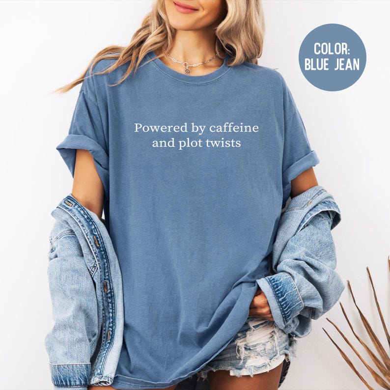 May include: A blue jean colored t-shirt with the text "Powered by caffeine and plot twists" in white. The shirt has rolled-up sleeves and a relaxed fit. A denim jacket is draped over the shoulders.