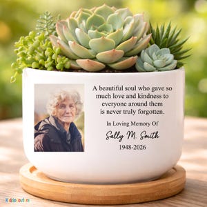 May include: White ceramic planter with a succulent arrangement, featuring a photo and text. The text reads "A beautiful soul... In Loving Memory Of Sally M. Smith 1948-2026". The planter sits on a wooden saucer, with a blurred green background.