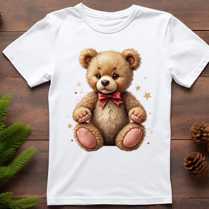 May include: White t-shirt featuring a cute teddy bear illustration. The bear is brown with a red bow tie and pink accents. Gold stars and red dots surround the bear. The shirt is on a wooden surface with pine cones and greenery.