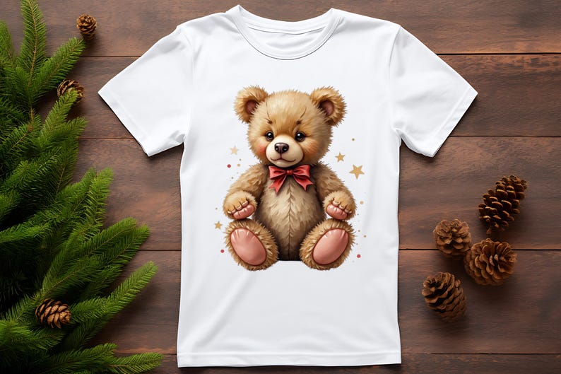 May include: White t-shirt featuring a cute teddy bear illustration. The bear is brown with a red bow tie and pink accents. Gold stars and red dots surround the bear. The shirt is on a wooden surface with pine cones and greenery.