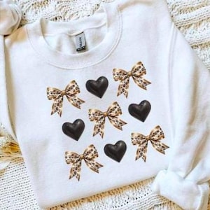 May include: White sweatshirt featuring a pattern of black hearts and leopard print bows. The bows have a tan and brown spotted design. The sweatshirt has a crew neck and long sleeves.