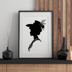 May include: Black and white silhouette print of a woman wearing a hat with feathers. The print is framed in a black frame and is displayed on a wooden shelf.