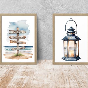 May include: Two framed wall art prints in light wood frames. The left print features a watercolor signpost with the text Cape Cod, Martha's Vineyard, Narragansett, and Portland on a beach. The right print shows a dark metal lantern with a lit candle.