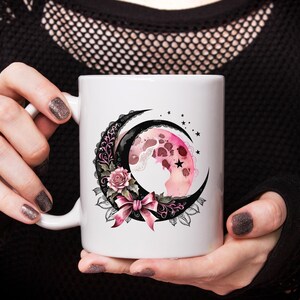 May include: White ceramic mug with a black and pink crescent moon design. The moon is decorated with a pink floral pattern and a black bow.