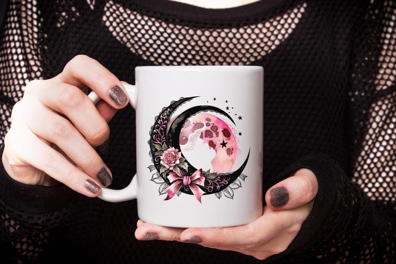 May include: White ceramic mug with a black and pink crescent moon design. The moon is decorated with a pink floral pattern and a black bow.