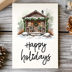 Puede incluir: Una tarjeta de felicitaci&oacute;n rectangular con una ilustraci&oacute;n de una caba&ntilde;a de madera marr&oacute;n decorada con guirnaldas verdes y una corona, rodeada de nieve. El texto Happy holidays est&aacute; impreso en cursiva negra bajo la ilustraci&oacute;n.