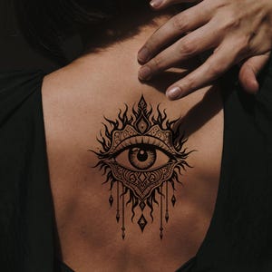 May include: A black tattoo of an eye with intricate ornamental details on a person's back. The eye is surrounded by flame-like shapes and decorative elements, with dangling teardrop-shaped pendants. The person is wearing a dark green top.