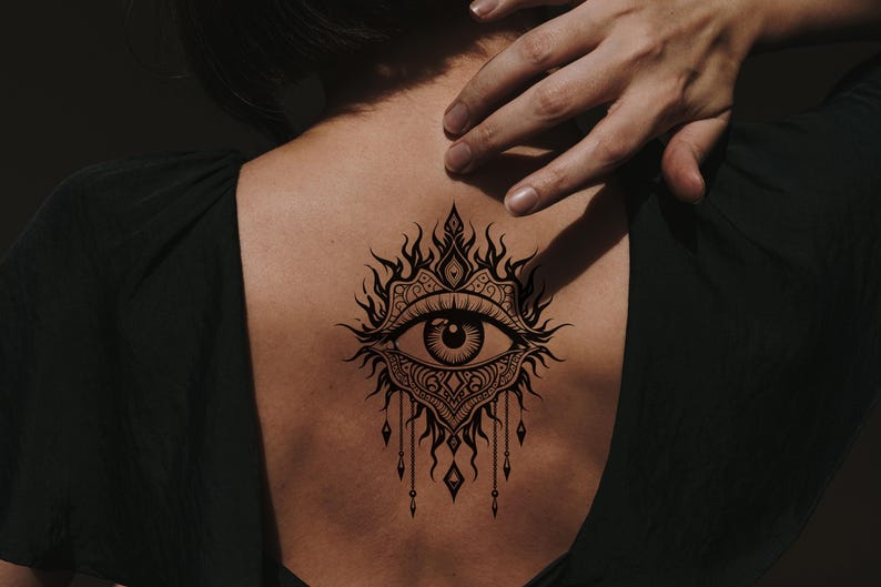 May include: A black tattoo of an eye with intricate ornamental details on a person's back. The eye is surrounded by flame-like shapes and decorative elements, with dangling teardrop-shaped pendants. The person is wearing a dark green top.