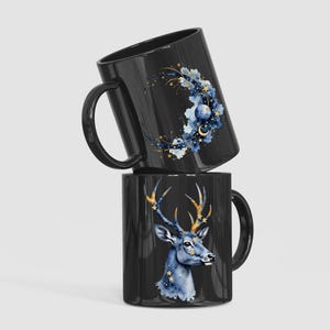 Op de afbeelding: Twee zwarte keramische mokken. E&eacute;n mok heeft een hemels ontwerp met een maan, sterren en bloemaccenten. De andere mok toont een aquarelillustratie van een hert met gouden geweien en sterdetails.