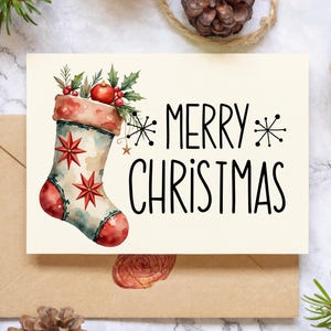 May include: A cream-colored Christmas card with a watercolor illustration of a festive stocking, adorned with red stars and berries. The card reads "MERRY CHRISTMAS" in black lettering. The card is placed on a marble surface with pinecones and greenery.