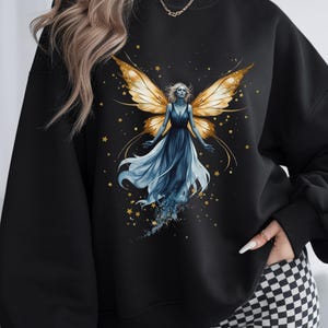 Op de afbeelding: Zwart sweatshirt met een aquarel illustratie van een fee met gouden vleugels en een vloeiende blauwe jurk. Het ontwerp bevat gouden sterren en een hemels thema, wat een grillige esthetiek cre&euml;ert.