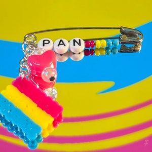 Puede incluir: Un imperdible con un dije adjunto. El dije es un corazón rosa con una cuenta blanca y una representación de cuentas de la bandera del orgullo pansexual. El imperdible tiene cuentas que deletrean "PAN" en él.