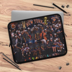 May include: A black laptop sleeve featuring a graphic print of the New York Knicks basketball team celebrating with championship trophies. The image includes the text NEW YORK in orange letters and the team logo in the upper left corner.