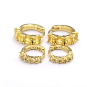 Op de afbeelding: Vier goudkleurige oorringen. Twee grotere ringen zijn versierd met rechthoekige gele edelstenen. Twee kleinere ringen hebben een rij kleinere, ronde gele edelstenen. De oorbellen staan op een witte achtergrond.