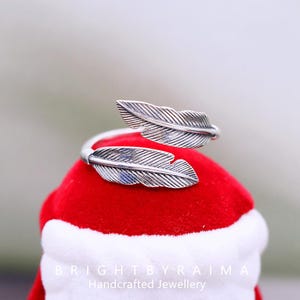 Può includere: Un anello a forma di piuma color argento. L'anello presenta due dettagliati disegni a forma di piuma che avvolgono il dito. È esposto su una superficie rossa con finiture bianche. Il testo "BRIGHTBYRAIMA Handcrafted Jewellery" è visibile.