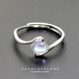 Könnte beinhalten: Ein silberner Ring mit einem ovalen, irisierenden Edelstein. Das Band des Rings hat ein fließendes Design, das den Stein umschließt. Der Ring wird auf dunklem Hintergrund präsentiert. Der Text "PAROLOVESHOP Handcrafted Jeweller" ist sichtbar.