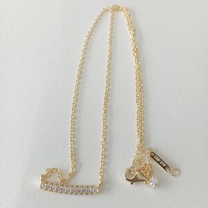 May include: A gold-colored necklace with a delicate chain and a bar pendant encrusted with small, clear gemstones. The necklace has a lobster clasp closure and a small tag with the text "ALE MET".
