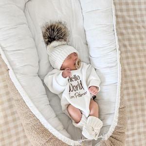 Może przedstawiać: Noworodek w białej dzianinowej czapce z dużym pomponem z futra, białym body z napisem "Hello World I'm Palmer" i pasującymi dzianinowymi bucikami. Dziecko leży w miękkim, beżowo-białym gnieździe dla dziecka.