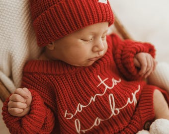 Puede incluir: Un bebé dormido con un conjunto rojo de punto de Santa Baby y un gorro a juego con un pompón blanco. El conjunto tiene las palabras "Santa Baby" en escritura blanca. El bebé está en una silla de madera.