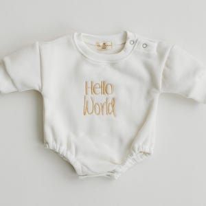 May include: Cream baby romper with short sleeves and a round neckline. The words "Hello World" are embroidered in gold thread on the front. The romper has snap closures on the shoulder and a gathered bottom.